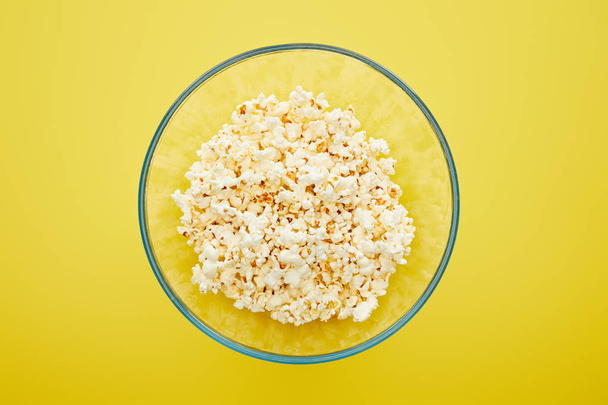 top view of delicious fresh popcorn in glass bowl on yellow background - Zdjęcie, obraz