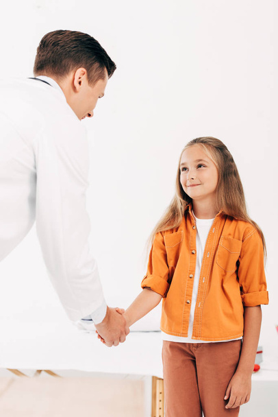 педиатр в белом халате и ребенок пожимают друг другу руки и смотрят друг на друга
 - Фото, изображение