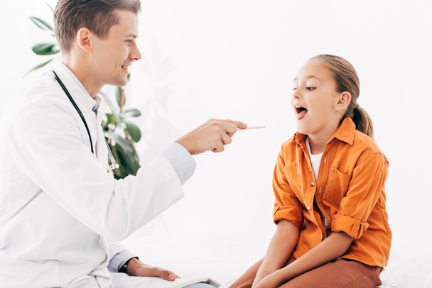 Kinderarzt im weißen Kittel untersucht Kind mit medizinischem Spachtel - Foto, Bild