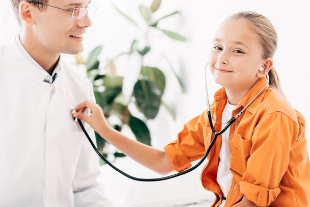 улыбающийся ребенок осматривает врача стетоскопом в клинике
 - Фото, изображение