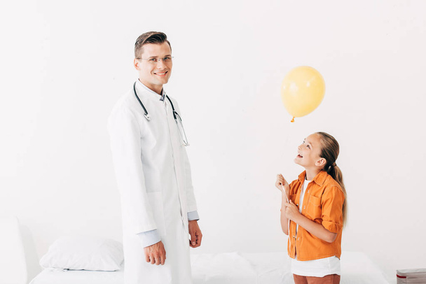 smiling pediatrist in white coat and happy child with balloon in clinic - Photo, Image