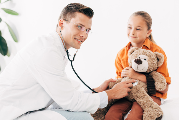 dziecko i uśmiechnięty rodak w białym płaszczu badanie misia z stetoskop w klinice - Zdjęcie, obraz