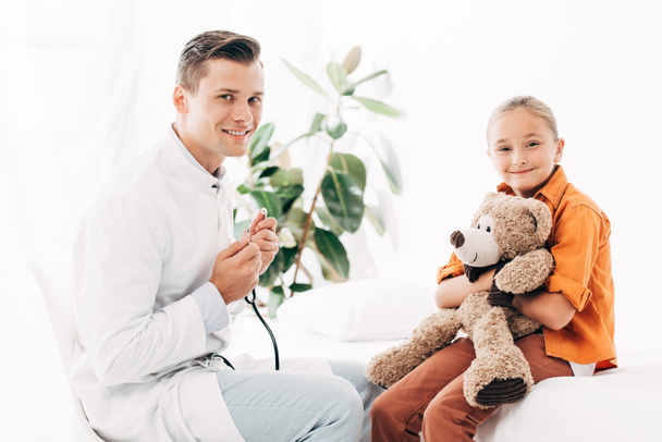 smiling pediatrist in white coat and kid with teddy bear in clinic - Foto, imagen