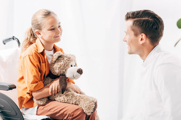 pediatrist and kid with teddy bear on wheelchair looking at each other - Photo, image