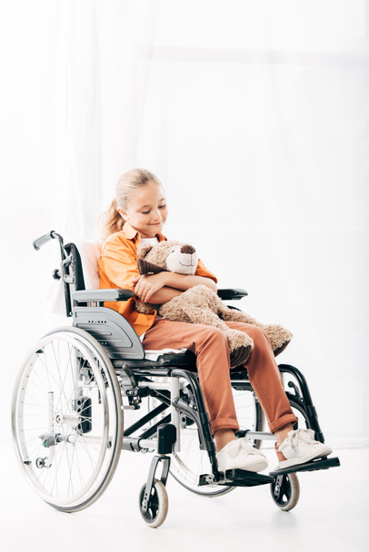 χαμογελαστός παιδί που κρατά το αρκουδάκι και κάθεται σε αναπηρικό καροτσάκι - Φωτογραφία, εικόνα