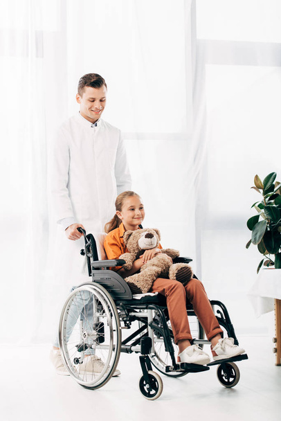 beyaz önlük ve tekerlekli sandalyede oyuncak ayı ile çocuk pediatrist tam uzunlukta görünümü - Fotoğraf, Görsel