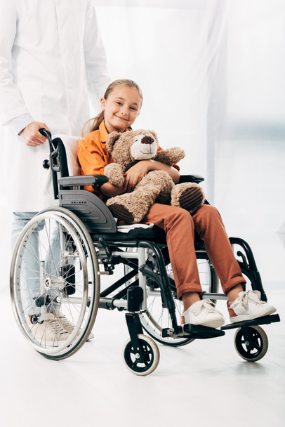 partial view of pediatrist in white coat and kid with teddy bear on wheelchair - Foto, afbeelding