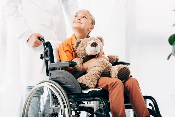 beyaz önlük ve tekerlekli sandalyede oyuncak ayı ile çocuk pediatrist kırpılmış görünümü - Fotoğraf, Görsel