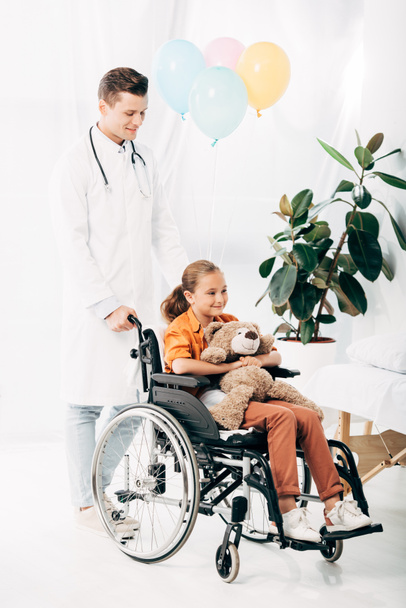 θέα πλήρους μήκους του pediatrist σε λευκό παλτό και παιδί με αρκουδάκι σε αναπηρικό καροτσάκι - Φωτογραφία, εικόνα