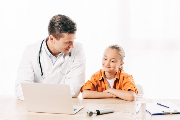 smiling pediatrist in white coat and child using laptop in clinic - Фото, зображення