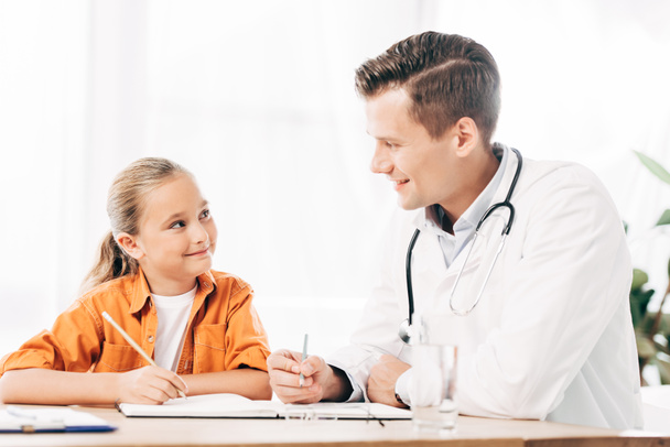 smiling child and pediatrist looking at each other while writing in notebook at table in clinic - Foto, imagen
