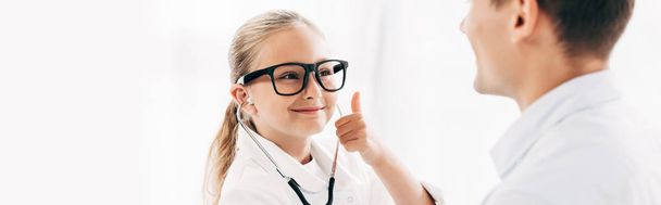 tiro panorâmico de criança sorridente em traje médico olhando para o paciente e mostrando o polegar para cima
 - Foto, Imagem