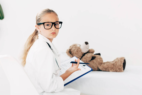 child in doctor costume holding clipboard and examining teddy bear - Zdjęcie, obraz