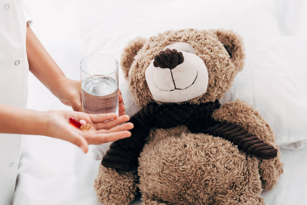 gedeeltelijke weergave van een kind dat glas water en pillen aan teddybeer geeft - Foto, afbeelding