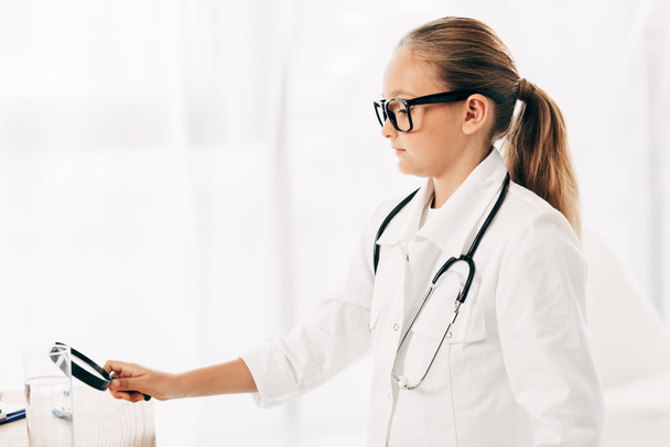 serious kid in doctor costume with stethoscope using magnifying glass - Zdjęcie, obraz