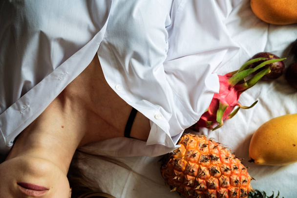 Young woman with long blonde hair wearing white home shirt lying on the bed. Summer healthy raw exotic fruits in bed. Top view, overhead. Time for relax and dream. Healthy eating concept - Foto, Bild