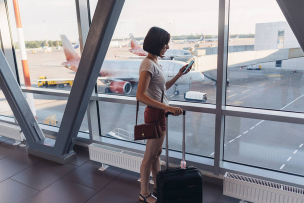 携帯電話を持って空港の窓の近くに立っている若い女性 - 写真・画像