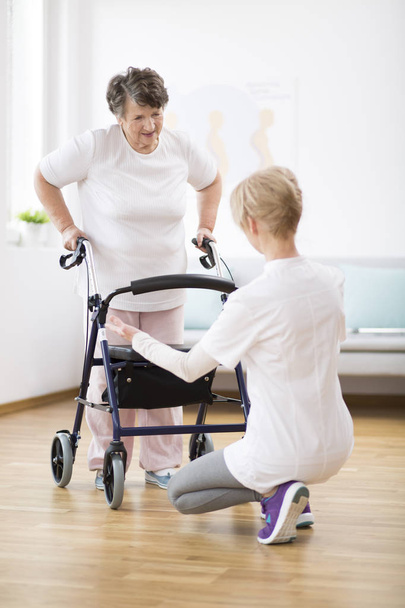 Senior vrouw met Walker probeert weer te lopen en behulpzame fysiotherapeut die haar steunt - Foto, afbeelding