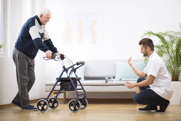 Elderly grandfather with walker trying to walk again and helpful male nurse supporting him - Foto, imagen