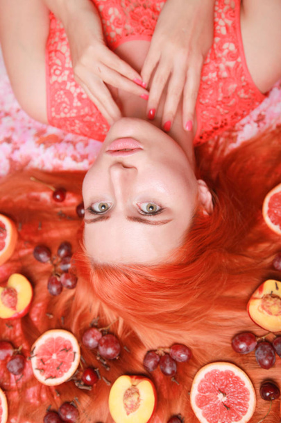 ピンクの明るい背景に彼女の髪の周りに果物で横たわっている美しい女性 - 写真・画像