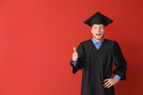 junger männlicher Student im Bachelor-Gewand zeigt Daumen nach oben auf farbigem Hintergrund - Foto, Bild
