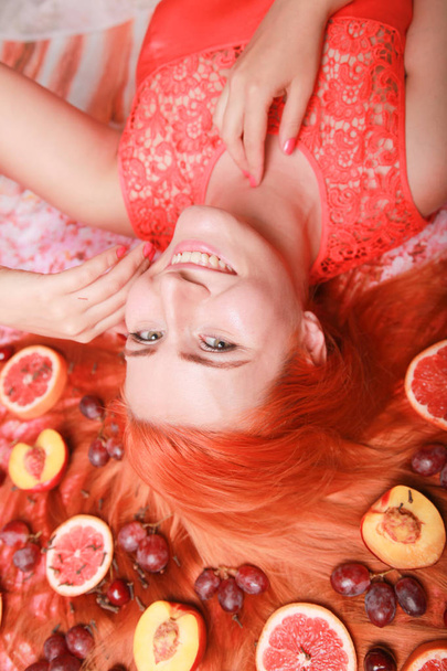 Beautiful woman lying with fruits around her hair on pink bright background - Photo, image