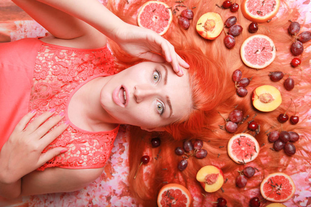 Beautiful woman lying with fruits around her hair on pink bright background - Foto, imagen
