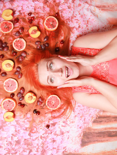 Beautiful woman lying with fruits around her hair on pink bright background - Photo, Image