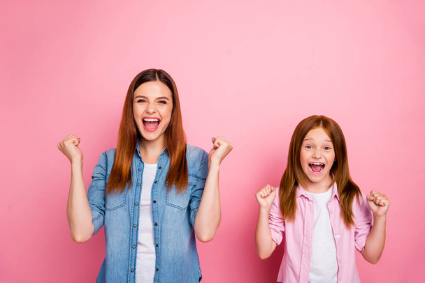Portrait of cheerful ladies with long hairdo raising fists screaming yeah wearing jeans denim shirts isolated over pink background - Photo, image