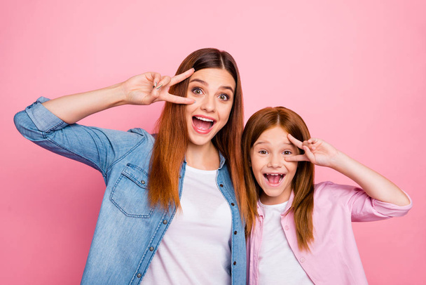 Portrait of cheerful ladies with long hair making v-signs near face wearing denim jeans shirt isolated over pink background - Фото, изображение