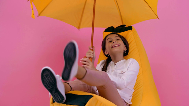 happy child relaxing with umbrella on bean bag chair isolated on pink - Footage, Video