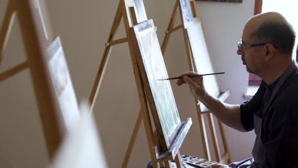 Artista masculino adulto en gafas pintando un cuadro usando pincel y pintura al óleo. Concepto artístico. Creativo pintor feliz pinta un cuadro colorido. Estudio de dibujo
 - Imágenes, Vídeo