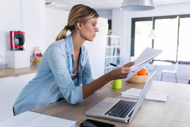 Pretty young woman working with laptop and documents in the kitchen at home. - 写真・画像