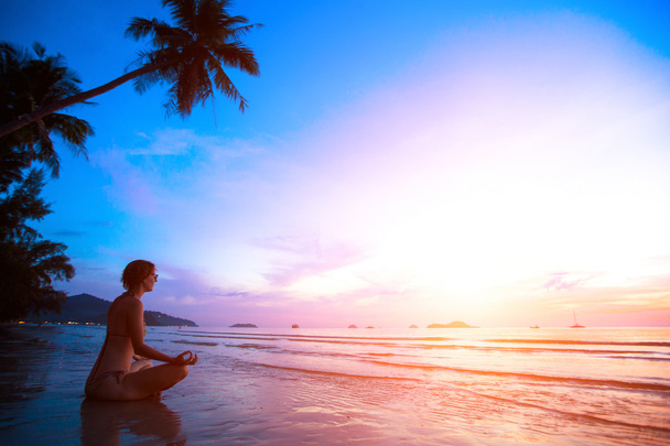 日没にビーチでヨガを練習している若い女性が - 写真・画像