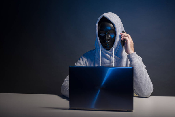 Anonimowy haker w masce programista używa laptopa i rozmawiać przez telefon, aby włamać się do systemu w ciemności. - Zdjęcie, obraz