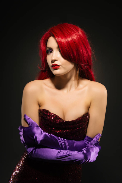Retrato de una atractiva mujer pelirroja con grandes pechos en un vestido de noche rojo brillante y guantes púrpura sobre un fondo oscuro
. - Foto, imagen