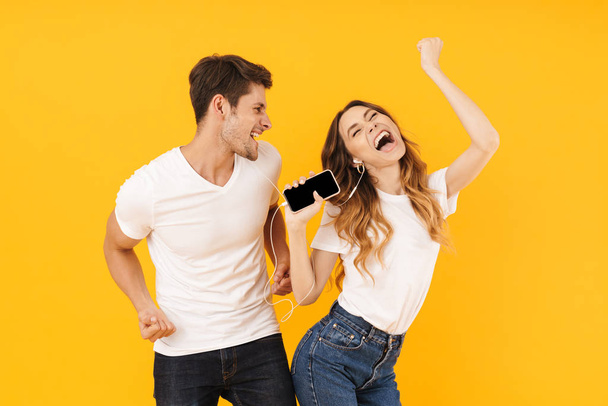 Portrait of joyful couple man and woman in basic t-shirts singing while using smartphone and earphones together - Foto, imagen