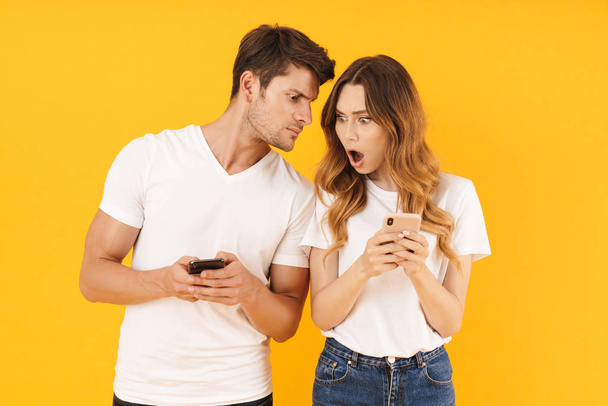 Foto van perplex paar man en vrouw in Basic t-shirts houden en gluren op mobiele telefoons - Foto, afbeelding
