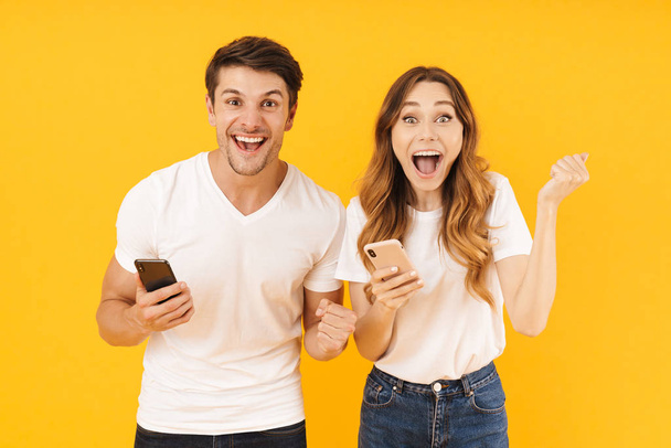 Portrait of happy couple man and woman in basic t-shirts rejoicing while standing together with smartphones - Foto, Bild