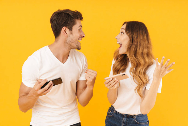 Portrait of cheery couple man and woman in basic t-shirts standing together while playing video games on smartphones - Foto, Bild