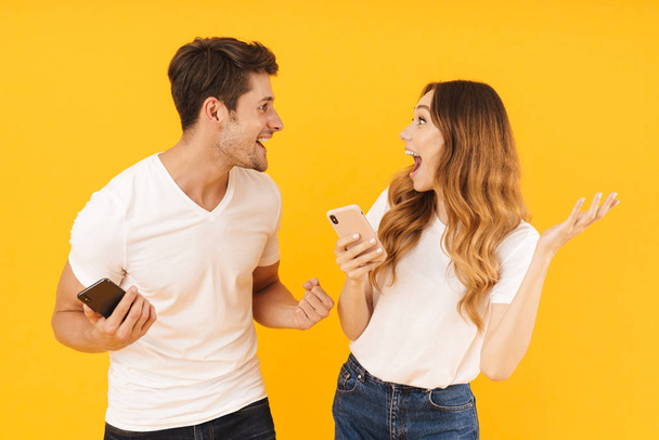 Portrait of attractive couple man and woman in basic t-shirts looking at each other while holding smartphones - Foto, Imagem