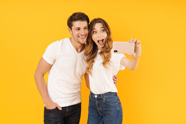 Portrait of astonished couple man and woman in basic t-shirts rejoicing while taking selfie photo together on smartphone - Foto, imagen
