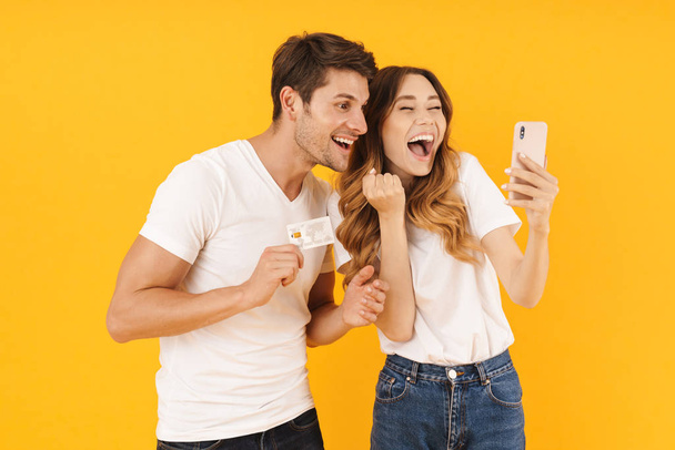 Portrait of caucasian couple in basic t-shirts rejoicing while holding credit card and smartphone - Φωτογραφία, εικόνα