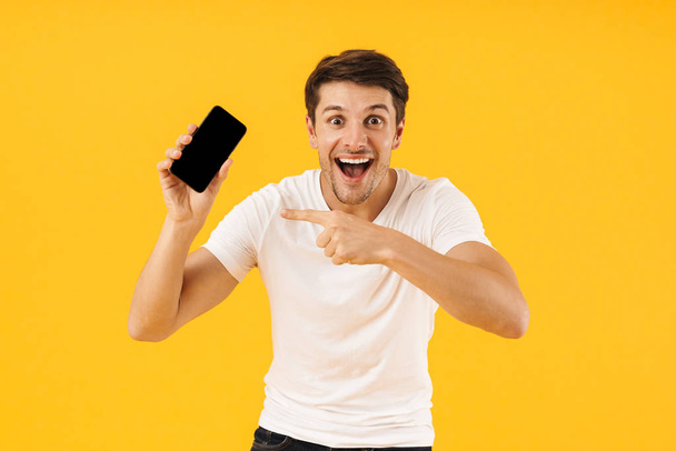 Shocked young man in casual white t-shirt using mobile phone isolated over yellow background. - Foto, Bild