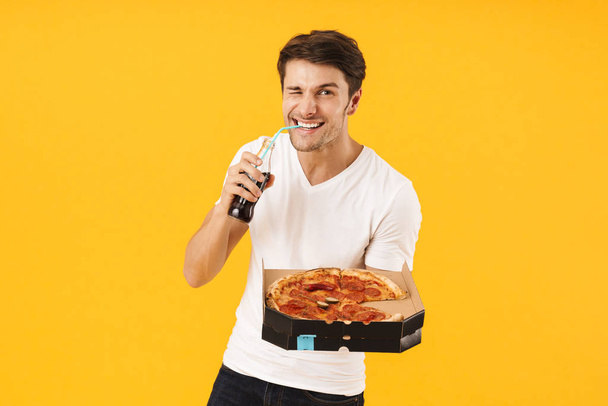 Cheerful happy young man in casual white t-shirt eat pizza drinking soda isolated over yellow background. - Фото, зображення