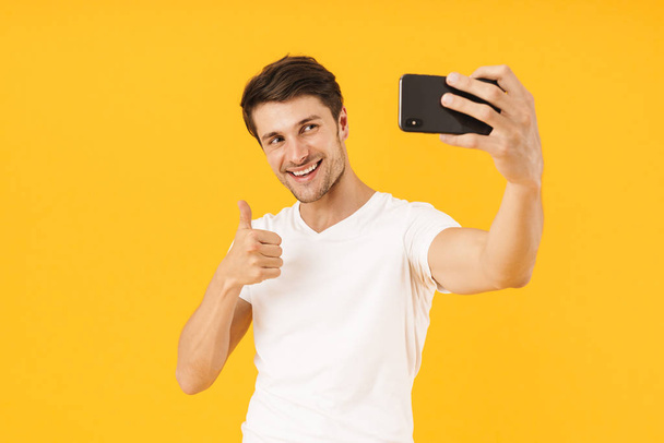 Happy young cheerful man in casual white t-shirt take a selfie by mobile phone isolated over yellow background. - 写真・画像