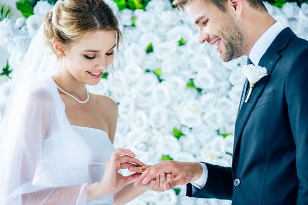 ελκυστική και χαμογελαστή νύφη βάζοντας βέρα γάμου στο δάχτυλο  - Φωτογραφία, εικόνα