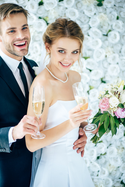 ελκυστική νύφη και όμορφος γαμπρός χαμογελώντας και κρατώντας γυαλιά σαμπάνιας - Φωτογραφία, εικόνα