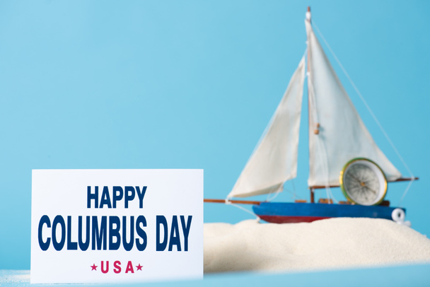 card with happy Columbus Day inscription near miniature ship in white sand isolated on blue - 写真・画像