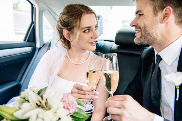 ελκυστική νύφη και όμορφος γαμπρός χαμογελά και κουδουνιά με ποτήρια σαμπάνιας - Φωτογραφία, εικόνα